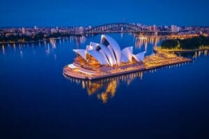 Aerial night view of the Sydney Opera House with its iconic white sails, surrounded by the illuminated waters of Sydney Harbour and the Harbour Bridge in the background.