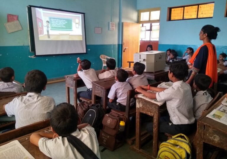 Classroom of young students in school uniforms attentively watching an educational video projected on a screen, with a teacher standing nearby guiding the lesson.
